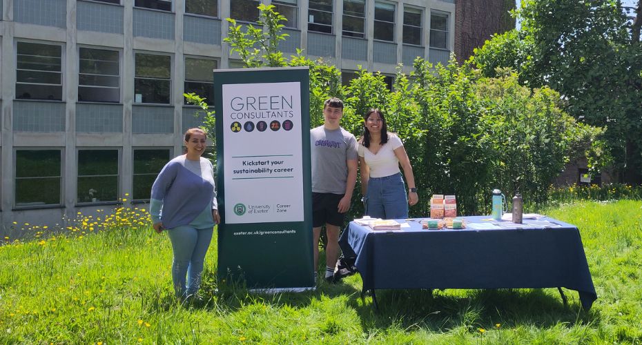 Three students stand in front of trees and next to a table displaying a Green Consultants sign,