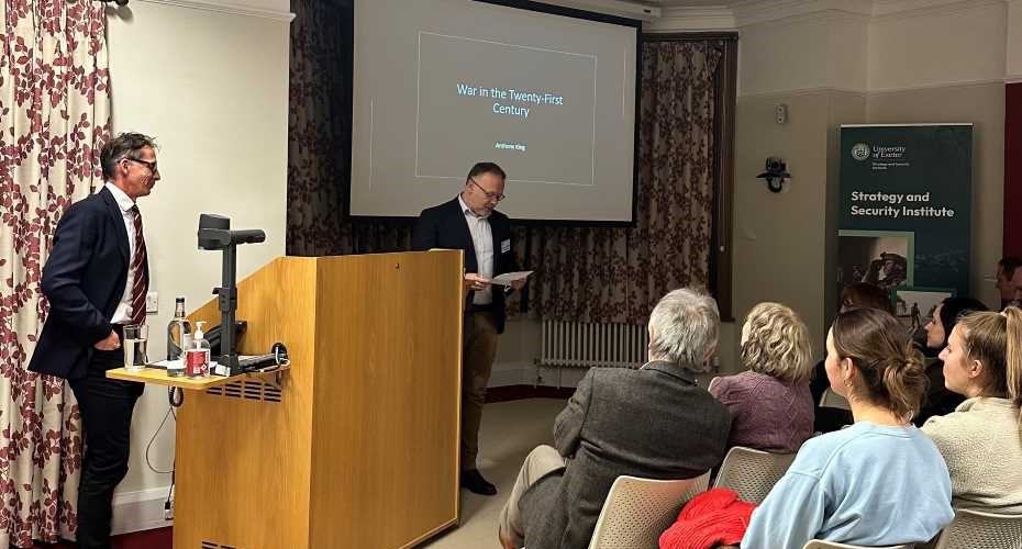A lecture room with two people stood beside a lecturn, a slide projection screen behind displaying the words 'War in the Twenty-First Century'. There is a banner saying Strategy and Security Institute. There is a number of people facing forward sat down attentively.