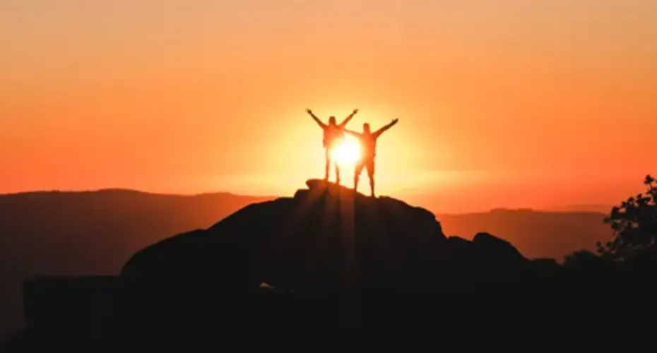 Silhouette image of two standing people on a mountain top at sunrise