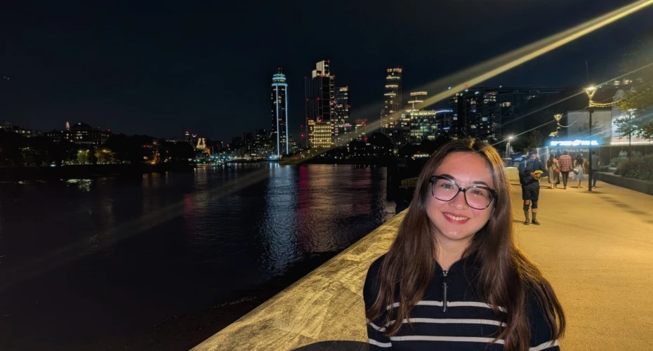 Profile image of Lucy Mitchell who has long hair, glasses and is smiling while stood in front of a river with tall buildings in the background.