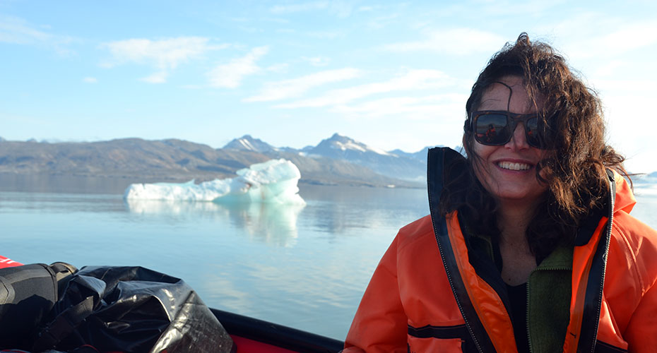 Portrait of Angela Gallego Sala in Svalbard, Norway. Angela looks windswept, but absolutely delighted. She is sitting in a small rowboat with a mass of ice floating behind. The sun is shining, but it looks very very cold.
