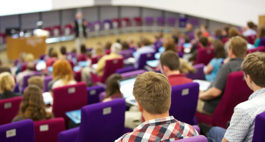 image taken from the back of a lecture whilst a lecture is taking place. You can see the backs of students head and in the distance there is a lecturer stood in front of an audience of students.