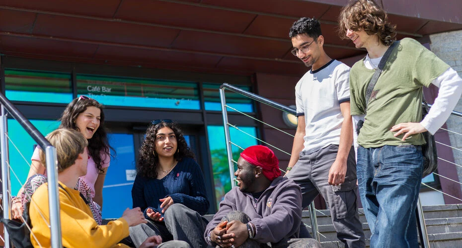 Image of a group of 6 students sat on steps outside of a building. Most can be seen smiling and laughing