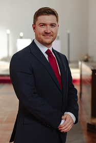 A photograph of Mr Michael Graham. He is wearing a red tie, white shirt and a dark blue suit, and is smiling at the camera. He is standing in the Mary Harris Memorial Chapel. The white east wall and altar, draped in a white altar-cloth, is softly focused behind him in the distance. The altar is partially sunlit, with silver-grey candlesticks gleaming in the sunlight.