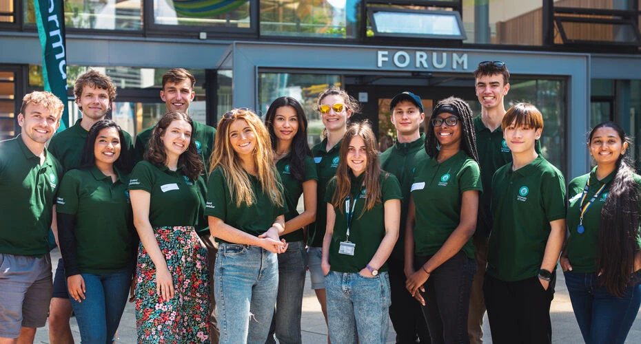 Image of a group of 14 students stood outside in front of the Forum building, all are wearing green exeter university tops